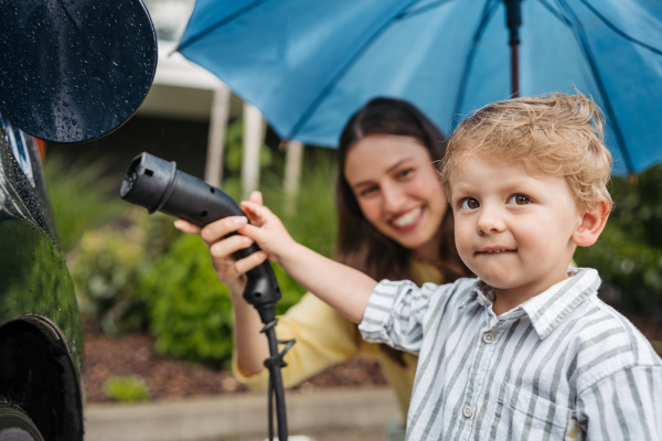 Smiling woman and her little son charging their electric car on a rainy day, standing under umbrella. Potential risks with charging an electric vehicle in the rain. Cute boy helping to plug in charger.