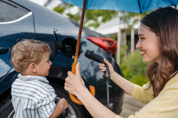 Smiling woman and her little son charging their electric car on a rainy day, standing under umbrella. Potential risks with charging an electric vehicle in the rain. Cute boy helping to plug in charger.