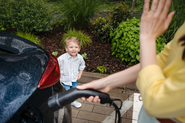 Cute little boy looking at his mother, while she charging electric car. Happy family in front house waiting to charge electric vehicle.