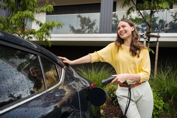 Young woman holding power supply cable from her car, prepared for charging it in home, sustainable and economic transportation concept.