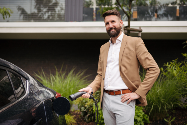 Portrait of businessman charging his electric car on the street. in front of his house. Pluging in charger in charging port.