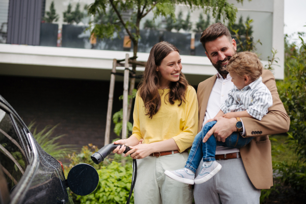 Happy family standing beside their electric car and charging it on the street. Electric vehicle with charger in charging port. Father holding son in his arms, showing him process of charging.