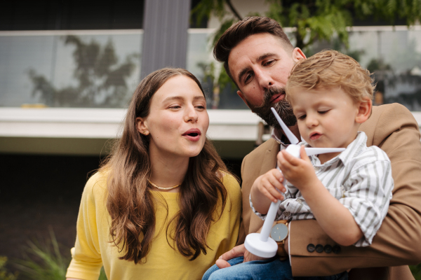 Smiling parents with little son blowing on wind turbine model. Teaching young children about renewable energy.