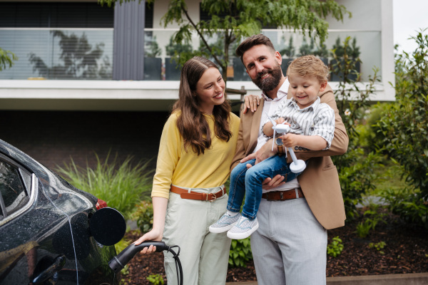 Happy family standing by beside their electric car and charging it on the street. Electric vehicle with charger in charging port.