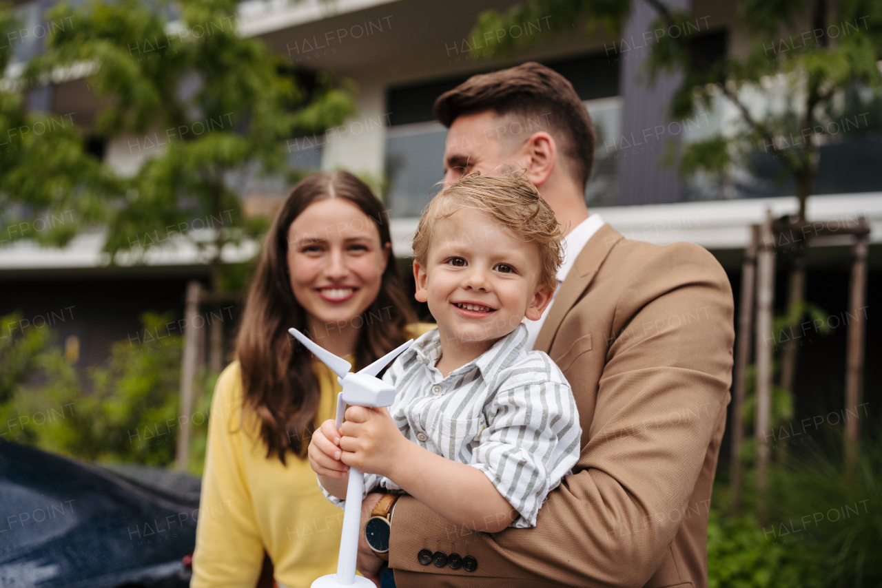 Portriat of cute boy holding wind turbine, while father charging their electric car on the street. Happy family in front house on rainy day.