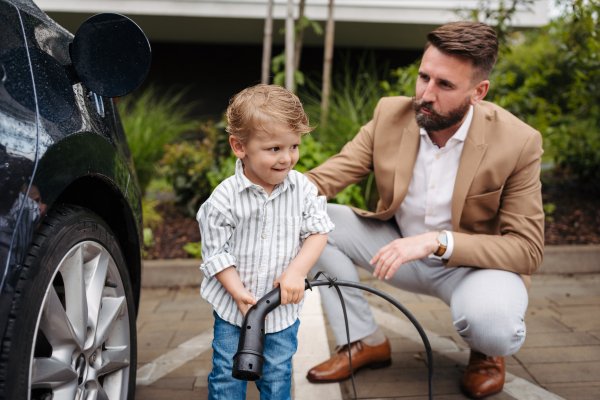 Smiling father and his little son charging electric car on the street. Cute boy helping his dad, learning how to plug in charger in charging port.