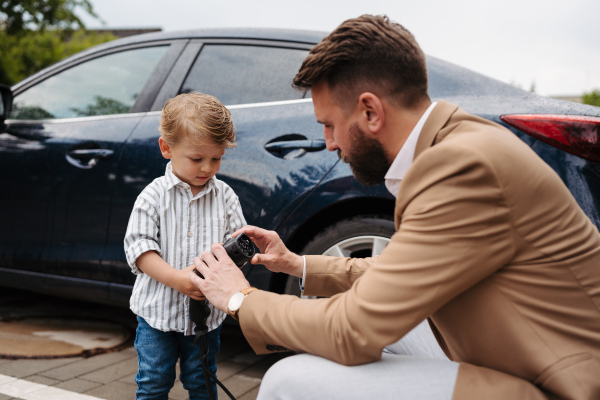 Father showing his son the electric vehicle charger. Mature man and his little son charging their electric car on the street. Cute little boy helping dad to plug in charger in charging port.