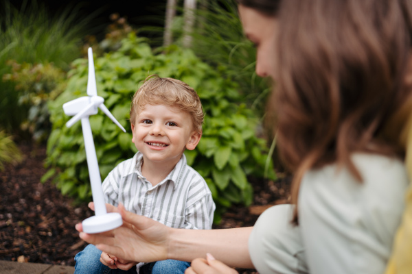 Portriat of cute boy holding wind turbine, while father charging their electric car on the street. Happy family in front house on rainy day.