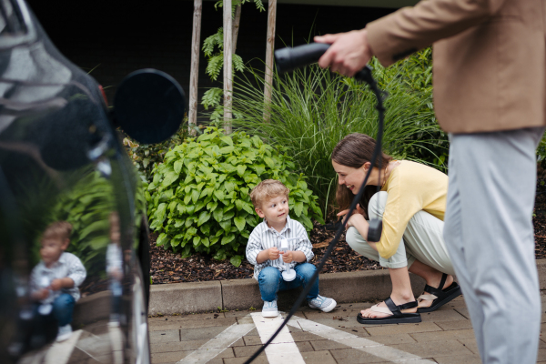 Man unplugging their electric car from charger on the street, while wife and son waiting for him. Happy family in front house waiting to charge electric vehicle.