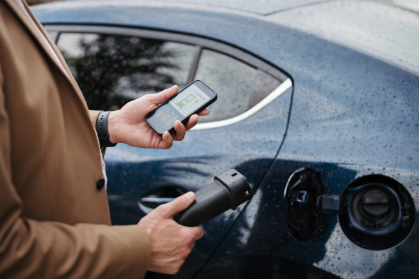 Close up of businessman using electric vehicle charging app, checking charging of his electric car from smart phone. Charging apps for monitoring electricity usage, locating charging stations.