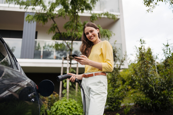 Smiling woman plugging charger in her electric car. Young businesswoman in front house waiting to charge electric vehicle.