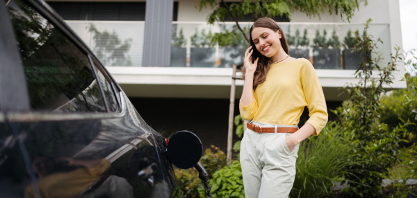 Close up of beautiful woman calling with smartphone while charging her electric car on the street in front of her house. Electric vehicle with charger in charging port. Banner with copy space.