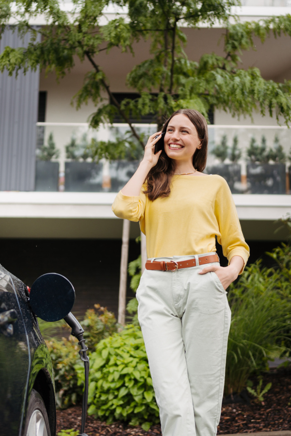 Close up of beautiful woman calling with smartphone while charging her electric car on the street in front of her house. Electric vehicle with charger in charging port. Banner with copy space.