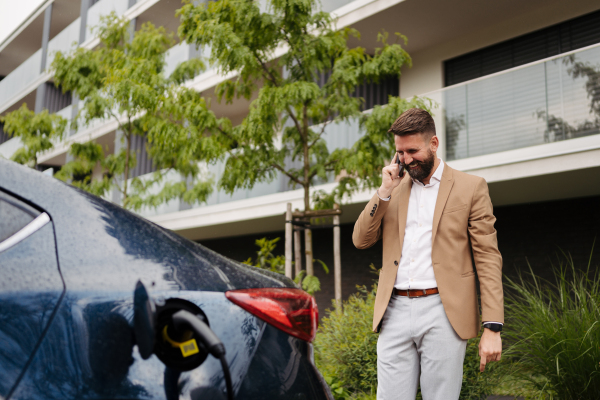 Close up of handsome businessman calling with smartphone while charging his electric car on the street in front of house. Electric vehicle with charger in charging port.