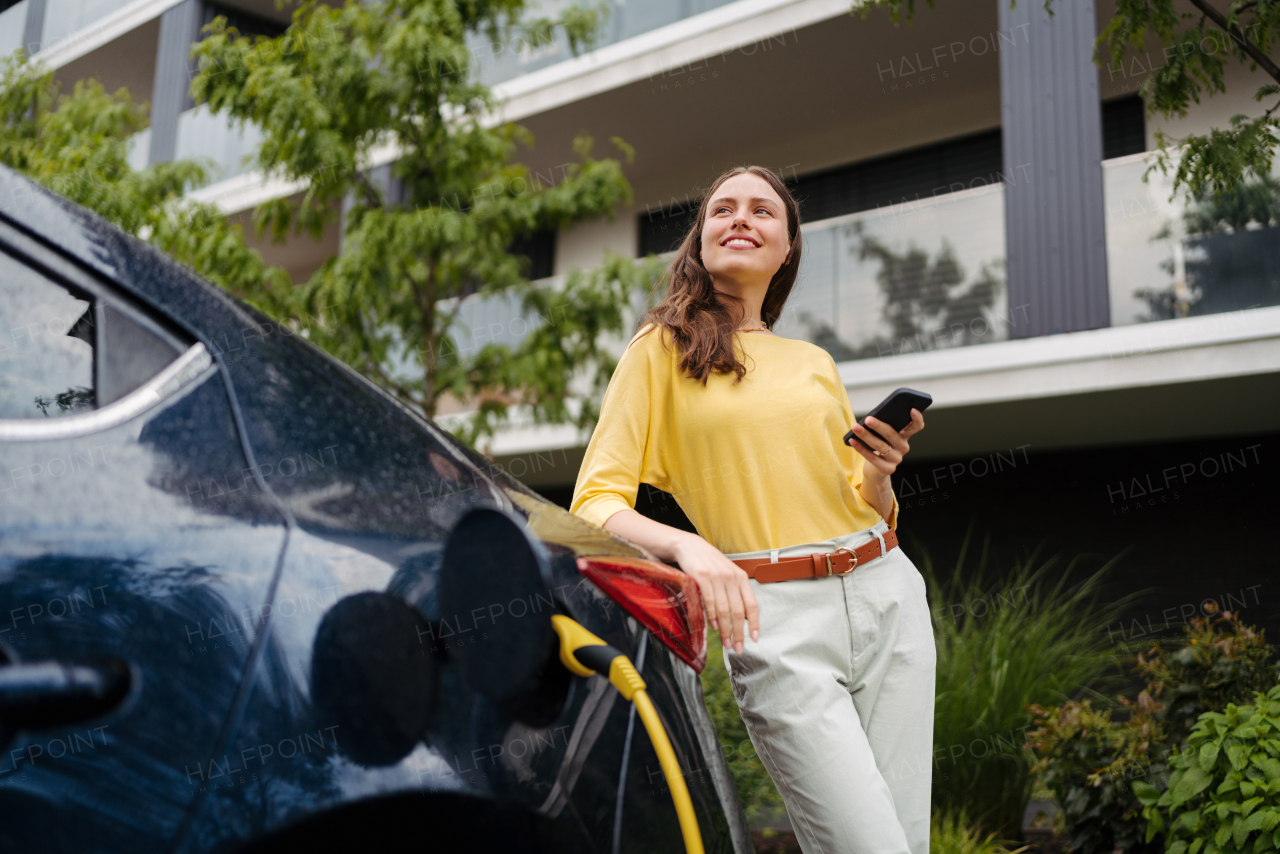 Close up of beautiful woman using smartphone while charging her electric car on the street in front of her house. Electric vehicle with charger in charging port.