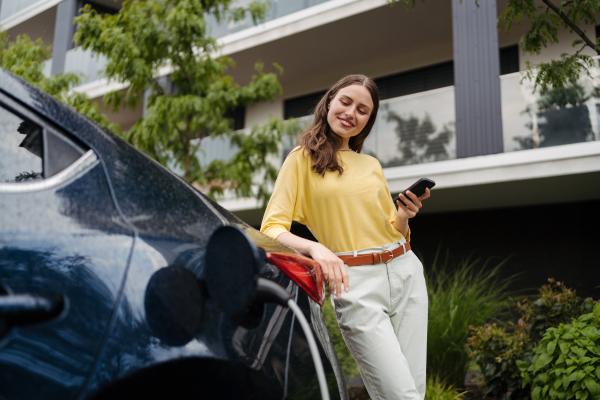 Smiling woman plugging charger in her electric car. Young businesswoman in front house waiting to charge electric vehicle. Female driver using electric vehicle charging app, checking progress of charging of electric car on smart phone.