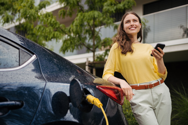 Young woman with smartphone waiting while electric car charging in home charging station, sustainable and economic transportation concept.