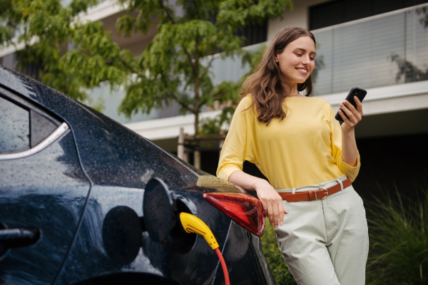 Close up of beautiful woman using smartphone while charging her electric car on the street in front of her house. Electric vehicle with charger in charging port.