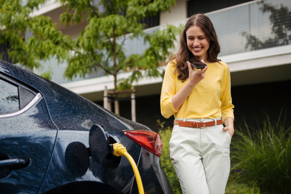 Close up of beautiful woman calling using speakerphone while charging her electric car on the street.r in front of her house. Electric vehicle with charger in charging port.