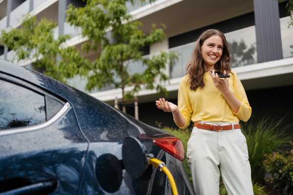 Close up of beautiful woman calling using speakerphone while charging her electric car on the street.r in front of her house. Electric vehicle with charger in charging port.