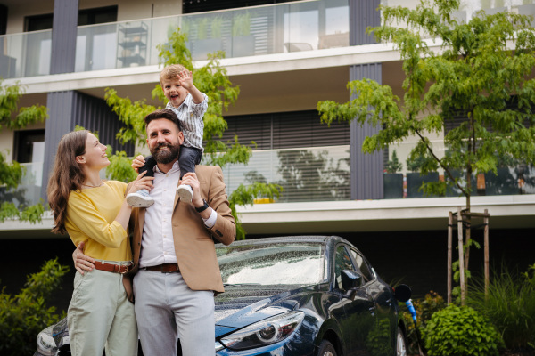 Happy family standing in front their electric car and charging it on the street. Electric vehicle with charger in charging port. Father carrying son on shoulders.