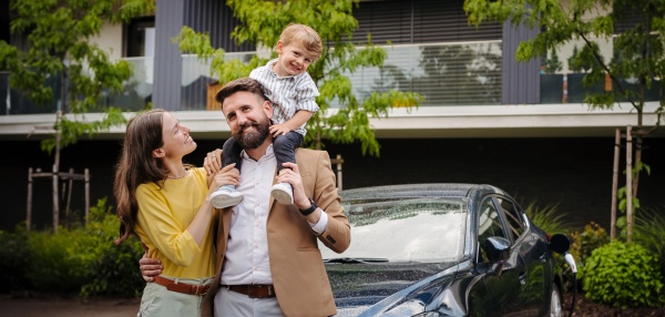 Happy family standing in front their electric car and charging it on the street. Electric vehicle with charger in charging port. Father carrying son on shoulders. Banner with coypa space.