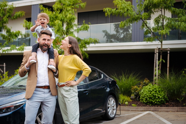 Happy family standing in front their electric car and charging it on the street. Electric vehicle with charger in charging port. Father carrying son on shoulders.