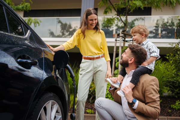 Happy family standing by beside their electric car and charging it on the street. Father holding son on his shoulders. Electric vehicle with charger in charging port.