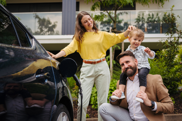 Happy family standing beside their electric car and charging it on the street. Electric vehicle with charger in charging port. Father holding son on his shoulders.