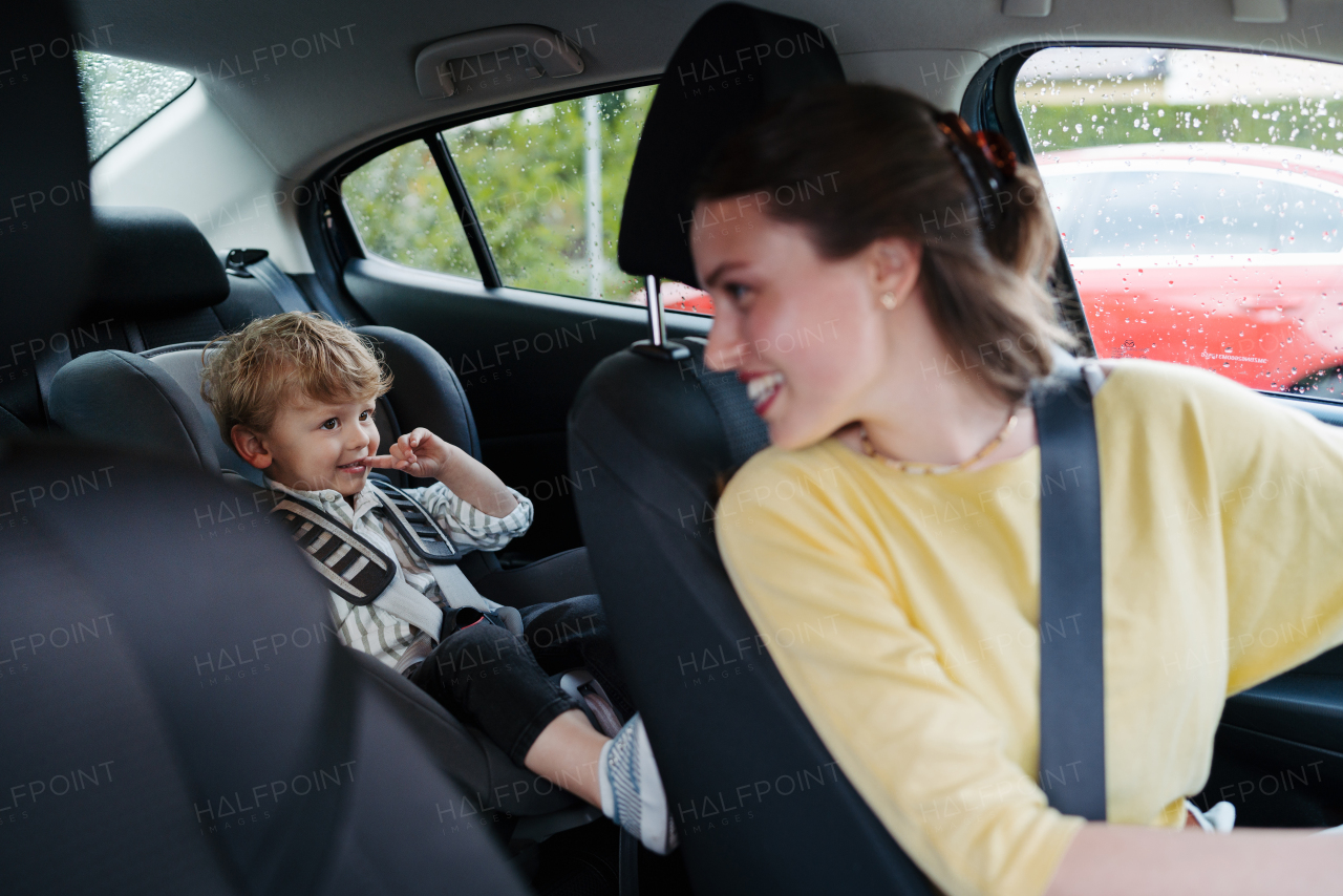 Cute little boy sitting in his car seat, playing on tablet. Mother checking her son before ride in car. Ride-on car safety. Driving in the rain.