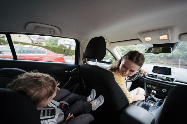 Cute little boy sitting in his car seat, playing on tablet. Mother checking her son before ride in car. Ride-on car safety. Driving in the rain.