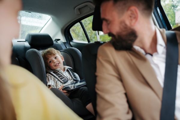 Cute little boy sitting in his car seat, playing on tablet. Parents checking son before ride in car. Ride-on car safety. Driving in the rain.