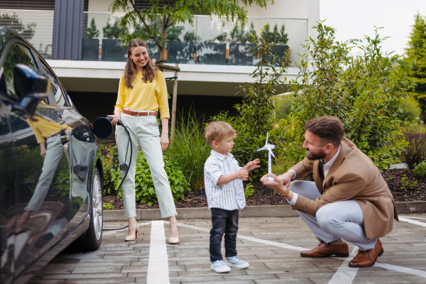 Happy family standing by beside their electric car and charging it on the street. Electric vehicle with charger in charging port. Little boy with dad playing with model of wind turbine.