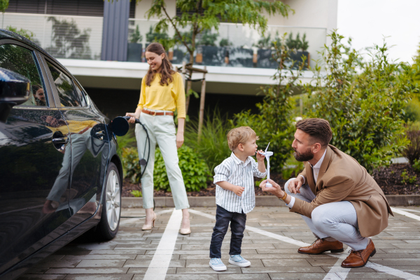 Portriat of woman charging electric car on the street, while husband and son waiting. Happy family in front house waiting to charge electric vehicle. Little boy blowing on wind turbine model. Teaching young children about renewable energy.