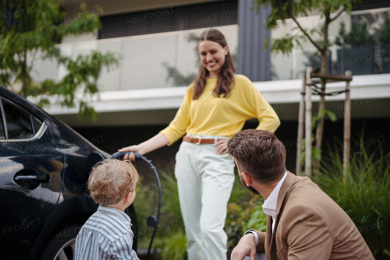 Portriat of woman charging electric car on the street, while husband and son looking at her. Happy family in front house waiting to charge electric vehicle.