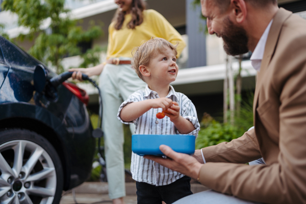 Son and father having snack while mother unplugging their electric car from charger. Happy family in front house waiting to charge electric vehicle.