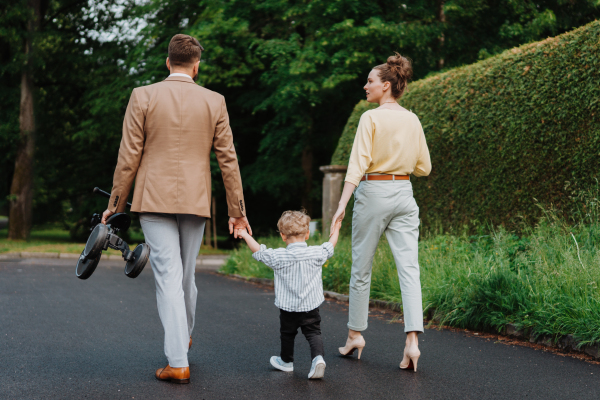 Rear view of young family with little son on a walk in a city park after work. Working parents spending time with their son after work day. Parents pick up kid from daycare.