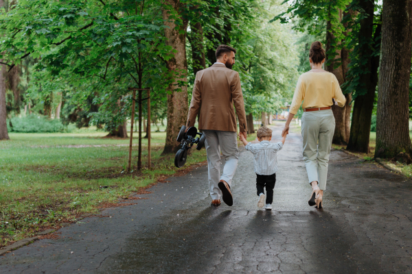 Rear view of young family with little son on a walk in a city park after work. Working parents spending time with their son after work day. Parents pick up kid from daycare.