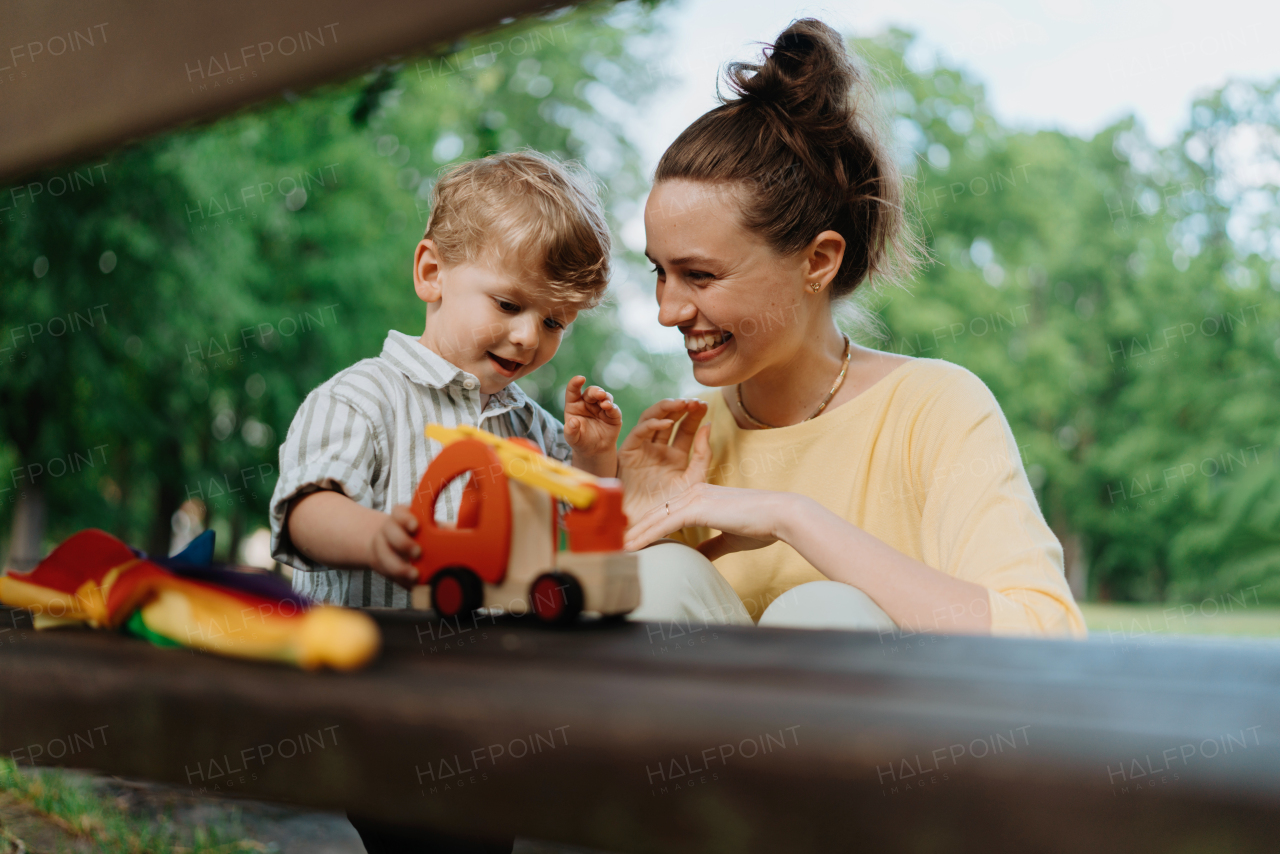 Single mother with little son playing with wooden toy on park bench. Working parent spending time with son in city park after work day.