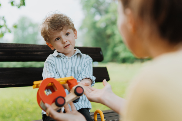 Single mother with little son playing with wooden toy on park bench. Working parent spending time with son in city park after work day.