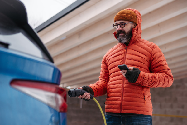 Mature man charging electric car during cold snowy day, using electric vehicle charging app, checking energy consumption, battery life on smart phone. Charging and driving electric vehicles during winter season.