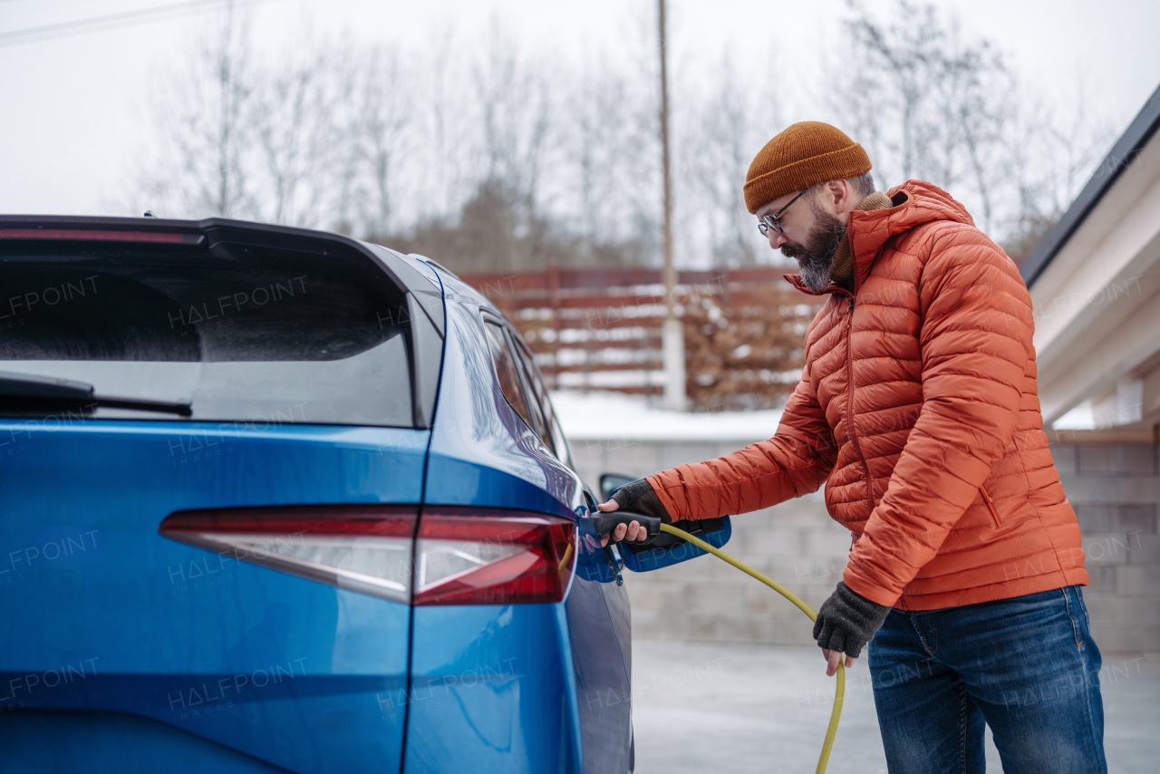 Man charging electric car during cold snowy day. Side view of hansome mature man putting charger in charging port during cold weather. Charging and driving electric vehicles during winter season.