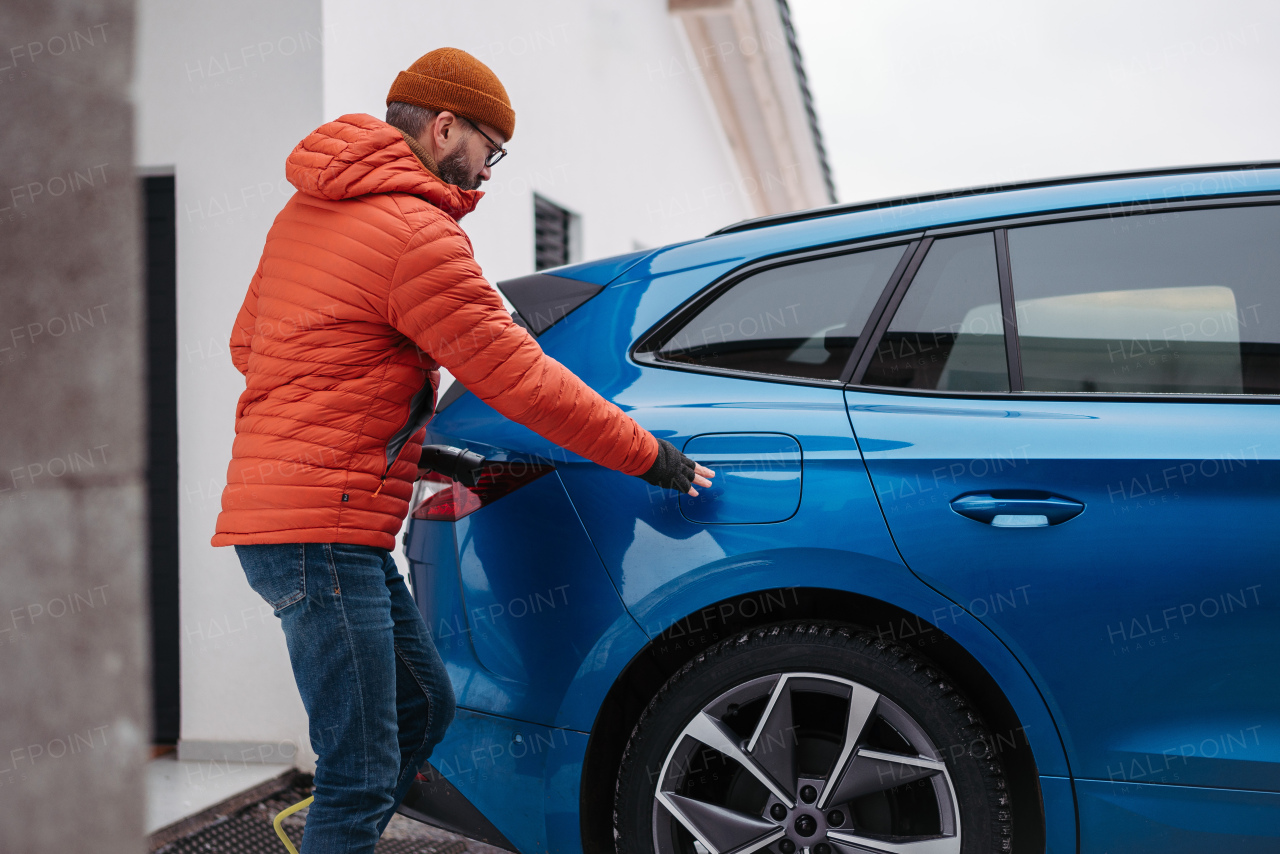 Man charging electric car during cold snowy day. Side view of hansome mature man putting charger in charging port during cold weather. Charging and driving electric vehicles during winter season.