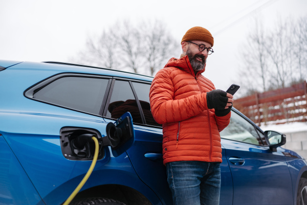 Mature man charging electric car during cold snowy day, using electric vehicle charging app, checking energy consumption, battery life on smart phone. Charging and driving electric vehicles during winter season.