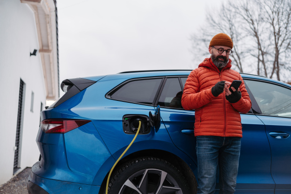 Mature man charging electric car during cold snowy day, using electric vehicle charging app, checking energy consumption, battery life on smart phone. Charging and driving electric vehicles during winter season.