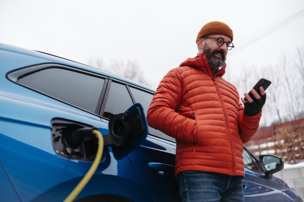 Mature man charging electric car during cold snowy day, using electric vehicle charging app, checking energy consumption, battery life on smart phone. Charging and driving electric vehicles during winter season.