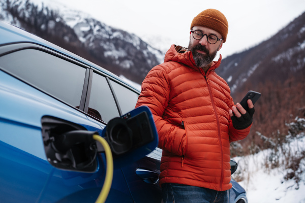 Mature man charging electric car during cold snowy day, using electric vehicle charging app, checking energy consumption, battery life on smart phone. Charging and driving electric vehicles during winter season.