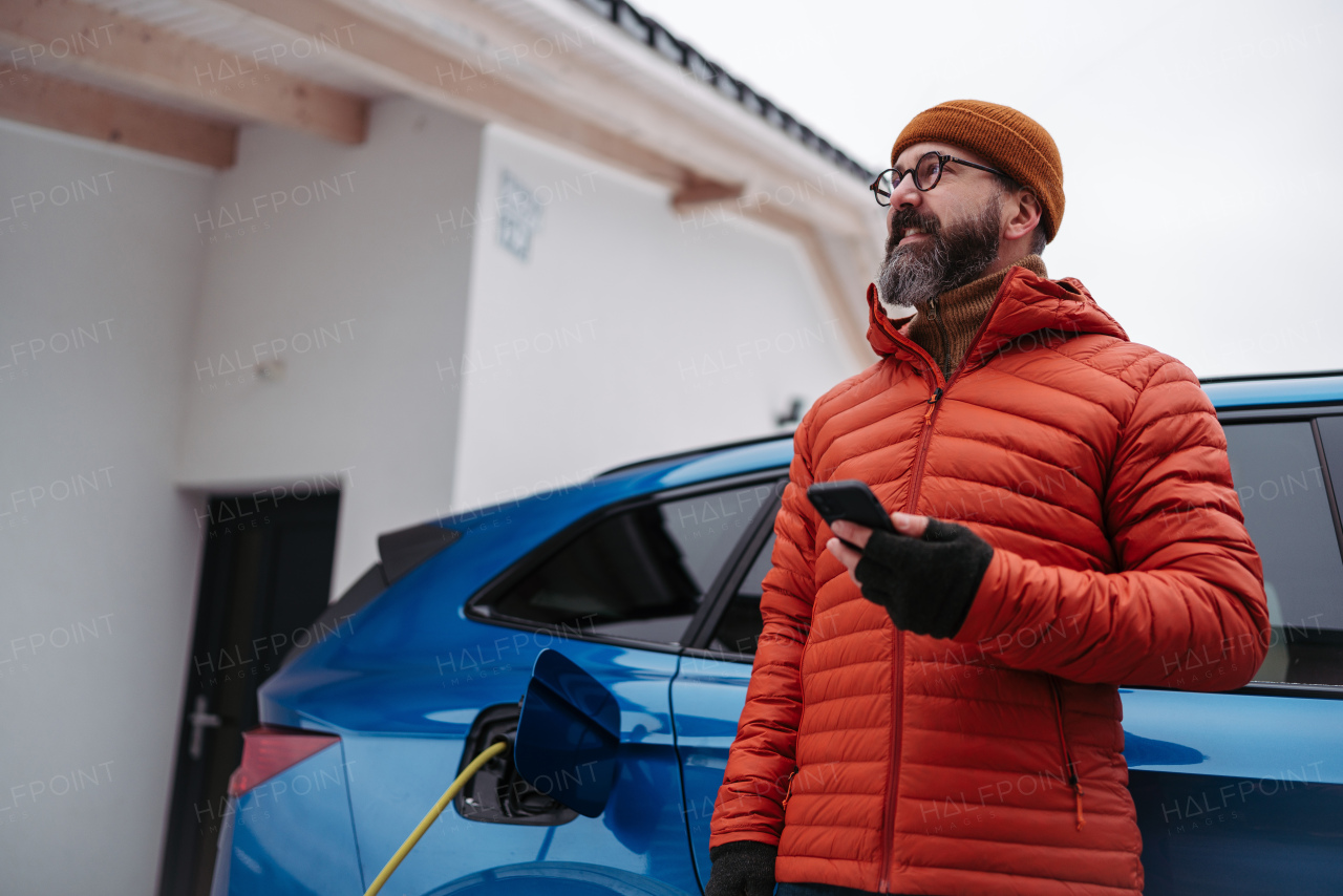 Mature man charging electric car during cold snowy day, using electric vehicle charging app, checking energy consumption, battery life on smart phone. Charging and driving electric vehicles during winter season.