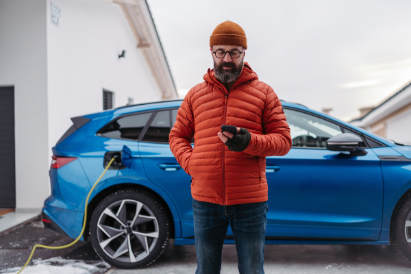 Mature man charging electric car during cold snowy day, using electric vehicle charging app, checking energy consumption, battery life on smart phone. Charging and driving electric vehicles during winter season.