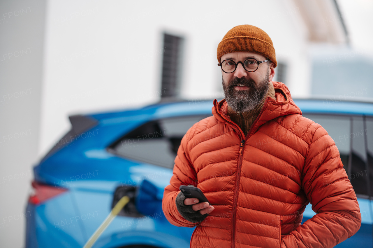 Mature man charging electric car during cold snowy day, using electric vehicle charging app, checking energy consumption, battery life on smart phone. Charging and driving electric vehicles during winter season.